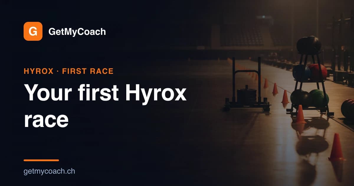 A loaded push sled and a rack of wall balls flanking an empty lane of polished floor in a floodlit indoor race arena, marker cones down the lane and empty grandstands behind — cover image for the GetMyCoach guide to your first Hyrox race.