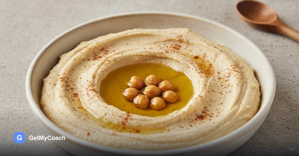 A shallow bowl of pale, very smooth hummus swirled into a well with a pool of olive oil, dusted with cumin and a few whole chickpeas on top.