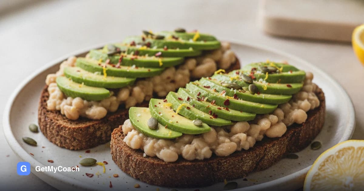 Two thick slices of toasted dark rye bread spread with coarsely crushed white beans and topped with sliced avocado, chilli flakes, lemon zest and pumpkin seeds.