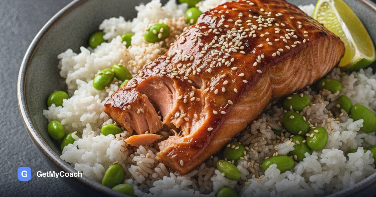 A bowl of rice topped with a glazed salmon fillet flaking apart, bright green edamame beans and a lime wedge, with sesame seeds scattered over.