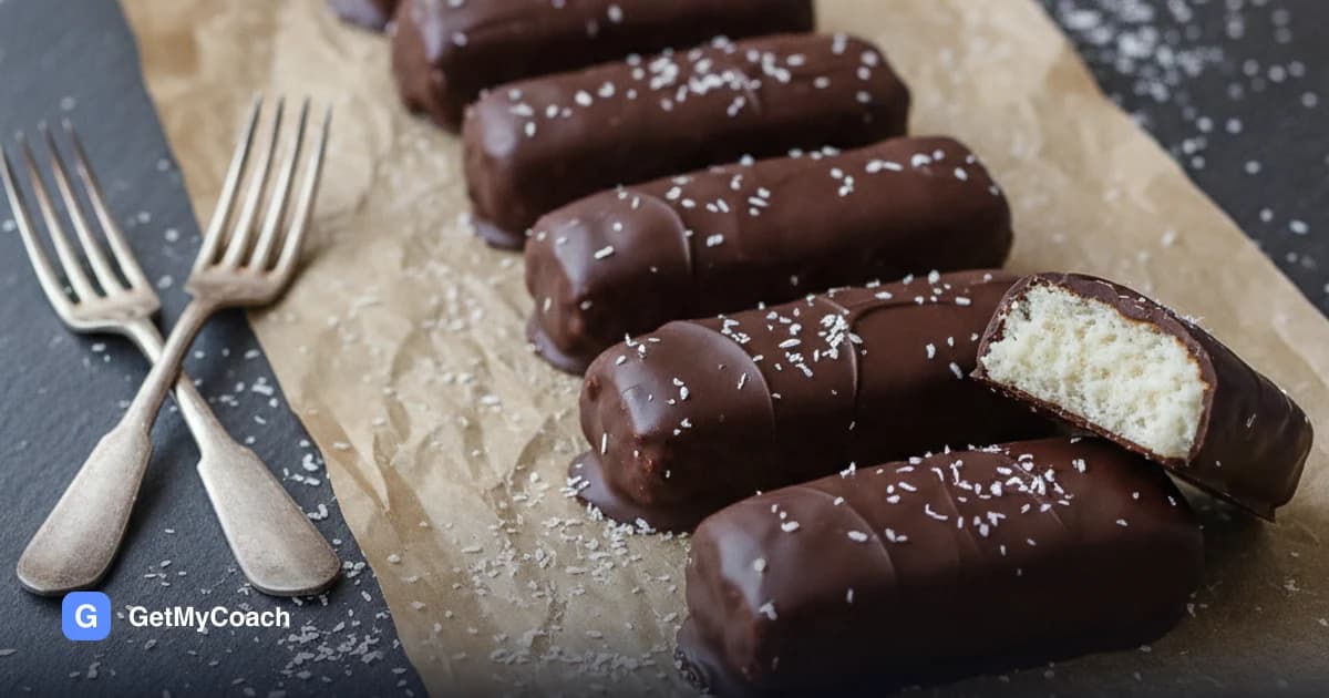 A row of coconut bars coated in dark chocolate on a sheet of crumpled baking parchment, one cut open to show the dense white coconut centre, with desiccated coconut scattered around them and two forks resting on the slate beside them.