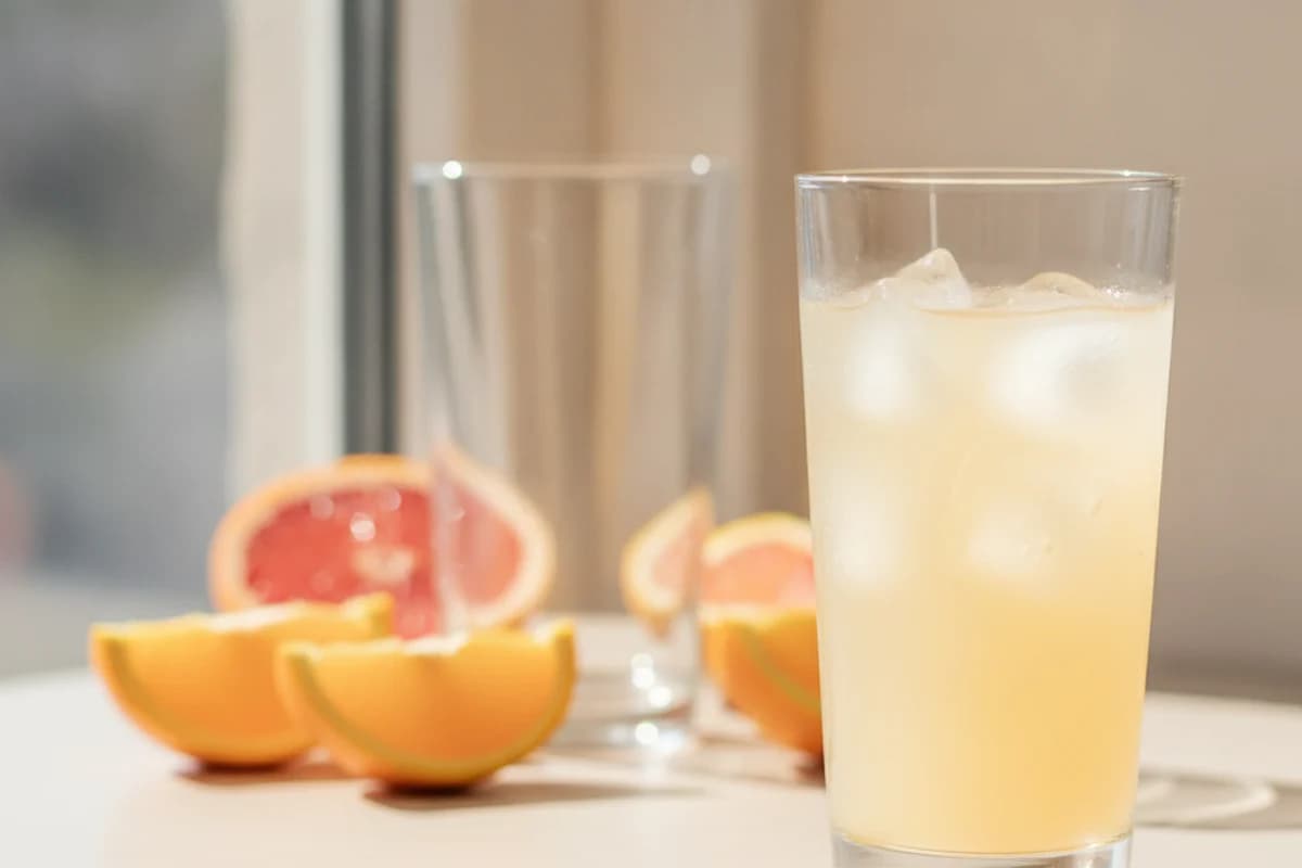 A tall clear glass of pale-orange fruit drink over large ice cubes with condensation on the outside, standing in bright daylight with a second empty glass and fruit slices out of focus behind.