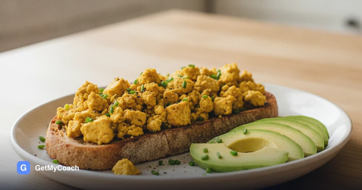 A thick slice of toasted sourdough topped with golden crumbled tofu flecked with black pepper and chives, slices of avocado fanned alongside, on a pale plate in morning light.