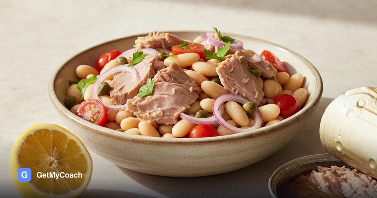 A bowl of white beans and flaked tuna with halved cherry tomatoes, capers, thin red onion and chopped parsley, a lemon half and an open tin beside it.